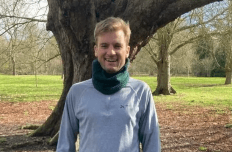 A man in a grey jumper stands in front of a tree smiling at the camera.