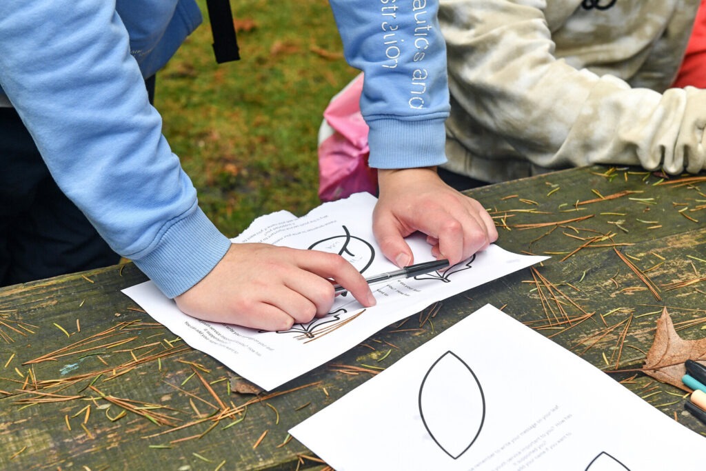 The hands of a young person sketching leaves on an outdoor table.