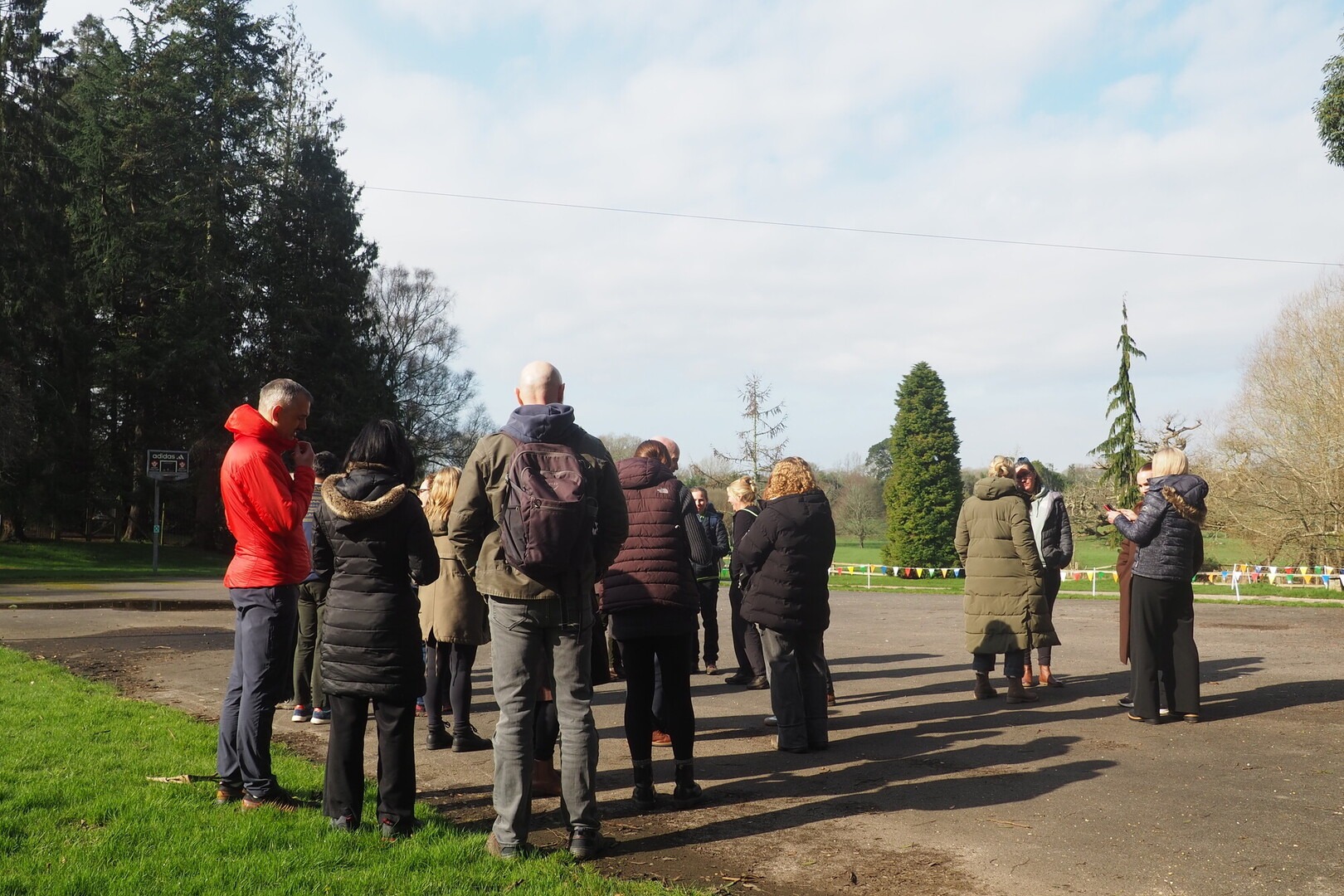 A group of people stand at the edge of a green field listening to a talk with their back to the camera.