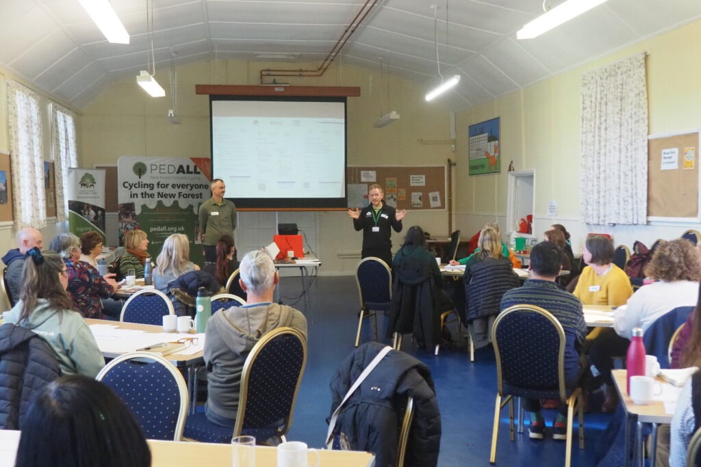 A room full of people take part in workshop discussions with a presenter standing at the front of the room beside a projector screen.