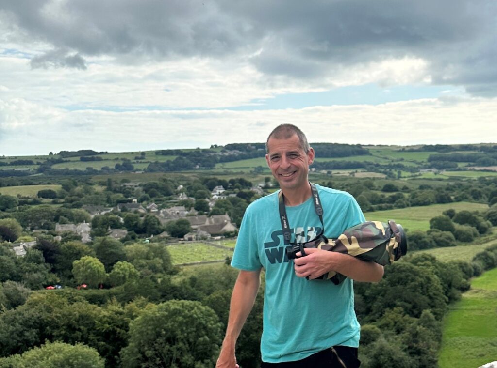 A man in a blue top stands holding a camera in front of a green landscape.