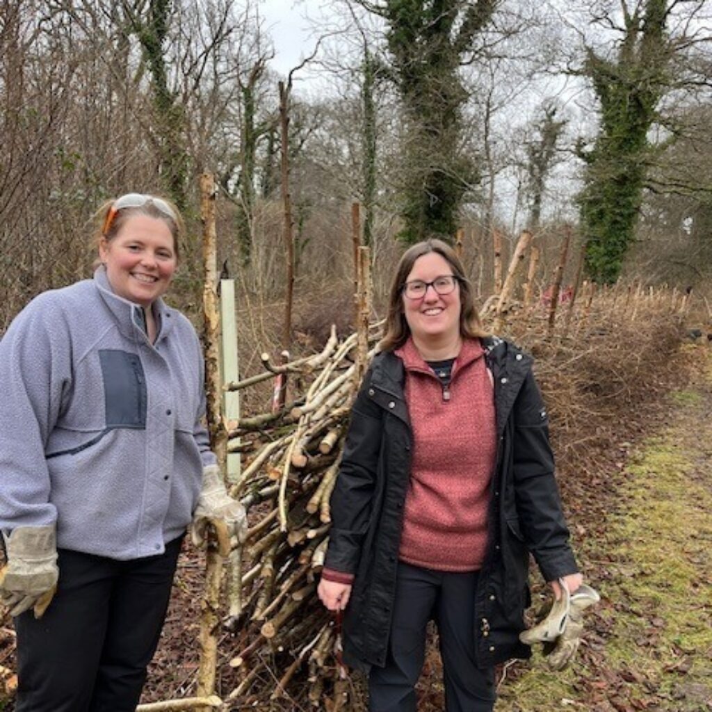 Two women smiling beside a stack of cut branches in a wooded area, one wearing gloves and one holding a face mask