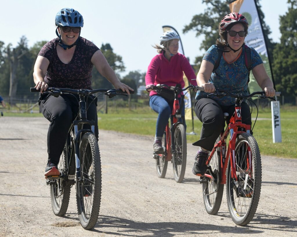 Three cyclists wearing helmets ride along a gravel path at an outdoor event, with two smiling riders in the foreground