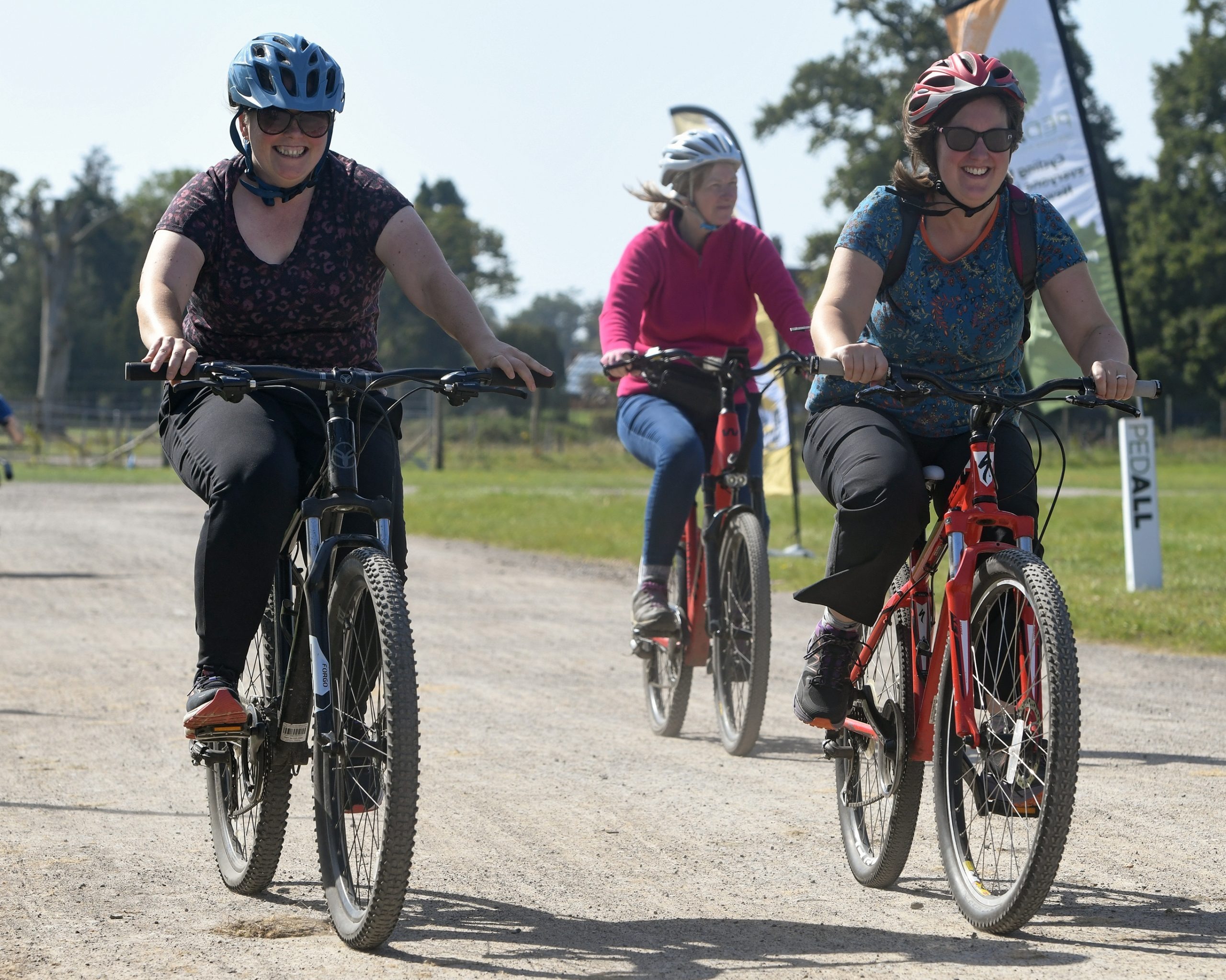 Three cyclists wearing helmets ride along a gravel path at an outdoor event, with two smiling riders in the foreground