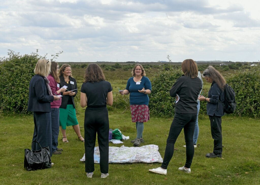 Group of people in a field listen to a speaker; map spread on the grass during an outdoor nature-connection activity