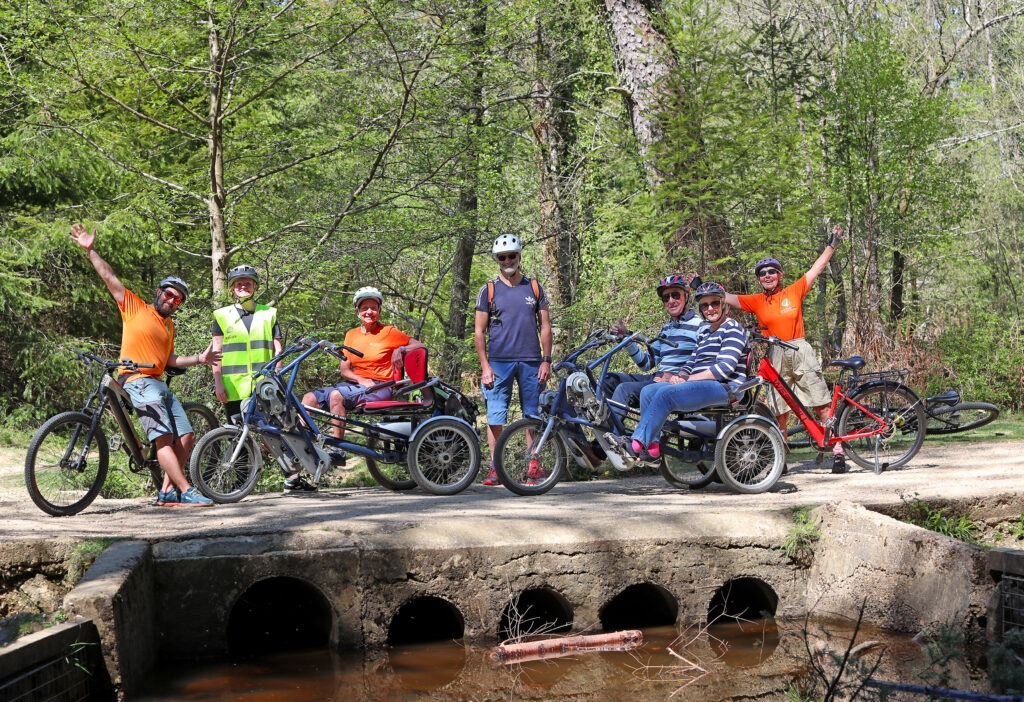 A group of riders on adapted cycles with PEDALL - New Forest Inclusive Cycling stop on a bridge over a stream in the New Forest, posing for the camera.