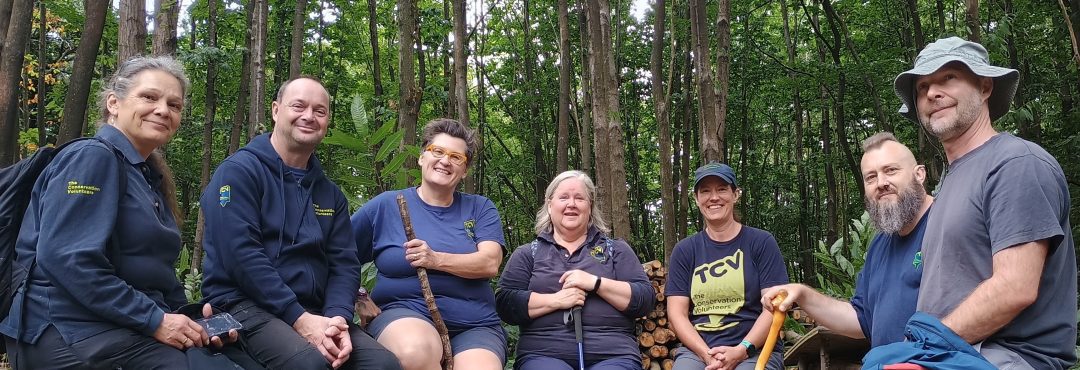 The Conservation Volunteers. a group of seven people sit in a woodland with trees behind them.