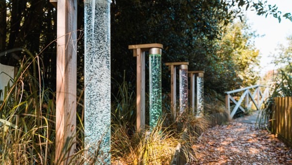 Garden path beside tall glass bubble columns in wooden frames, with grasses and a white gate in the background
